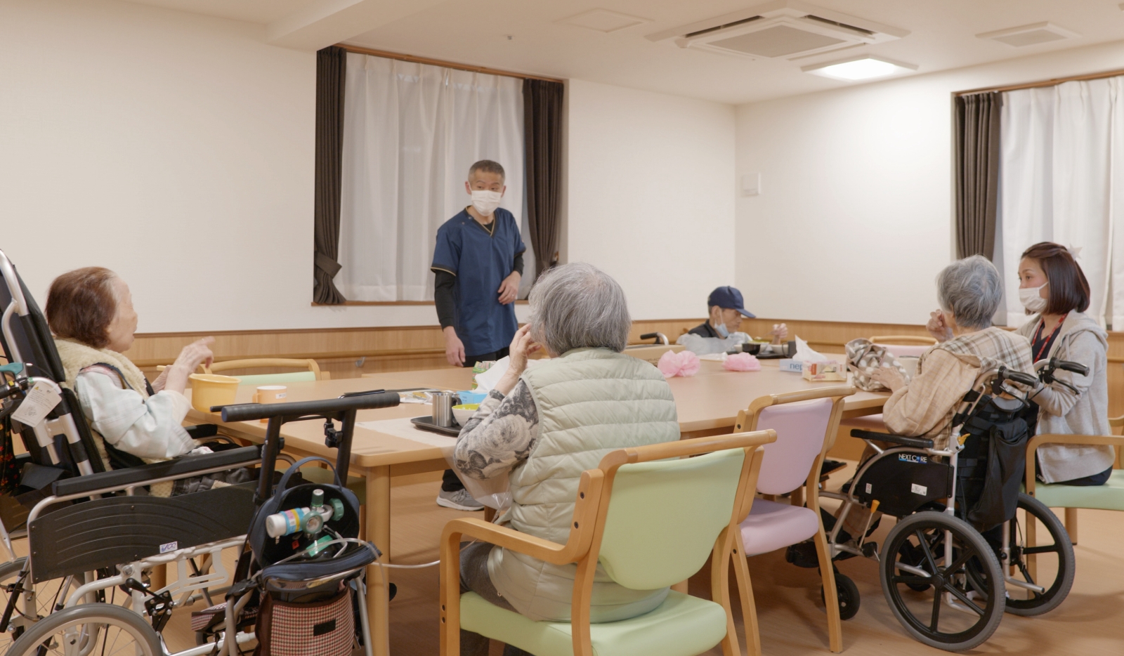 介護施設の共用スペースで職員が高齢者と交流する風景。レク活動や日中プログラム中の様子。